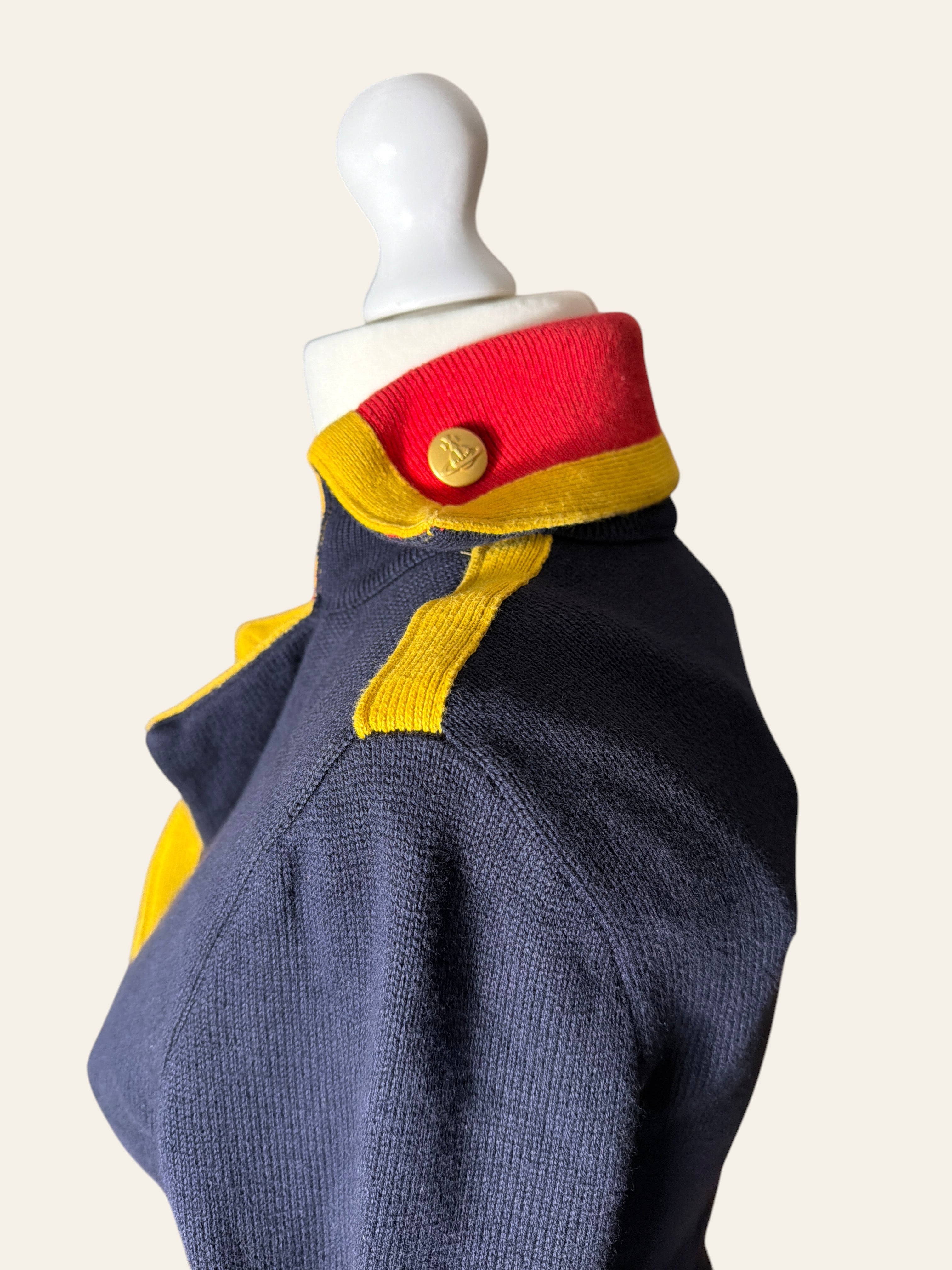 A close-up side view of a navy blue knit garment displayed on a white mannequin torso, featuring a high red and yellow collar with a gold button and yellow shoulder detailing, against a light beige background.