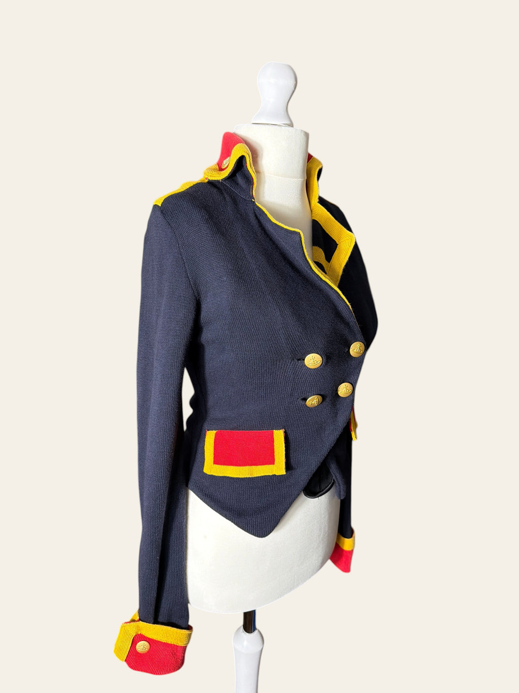 A navy blue military-style jacket with yellow and red trim, displayed on a white mannequin torso against a light beige background.