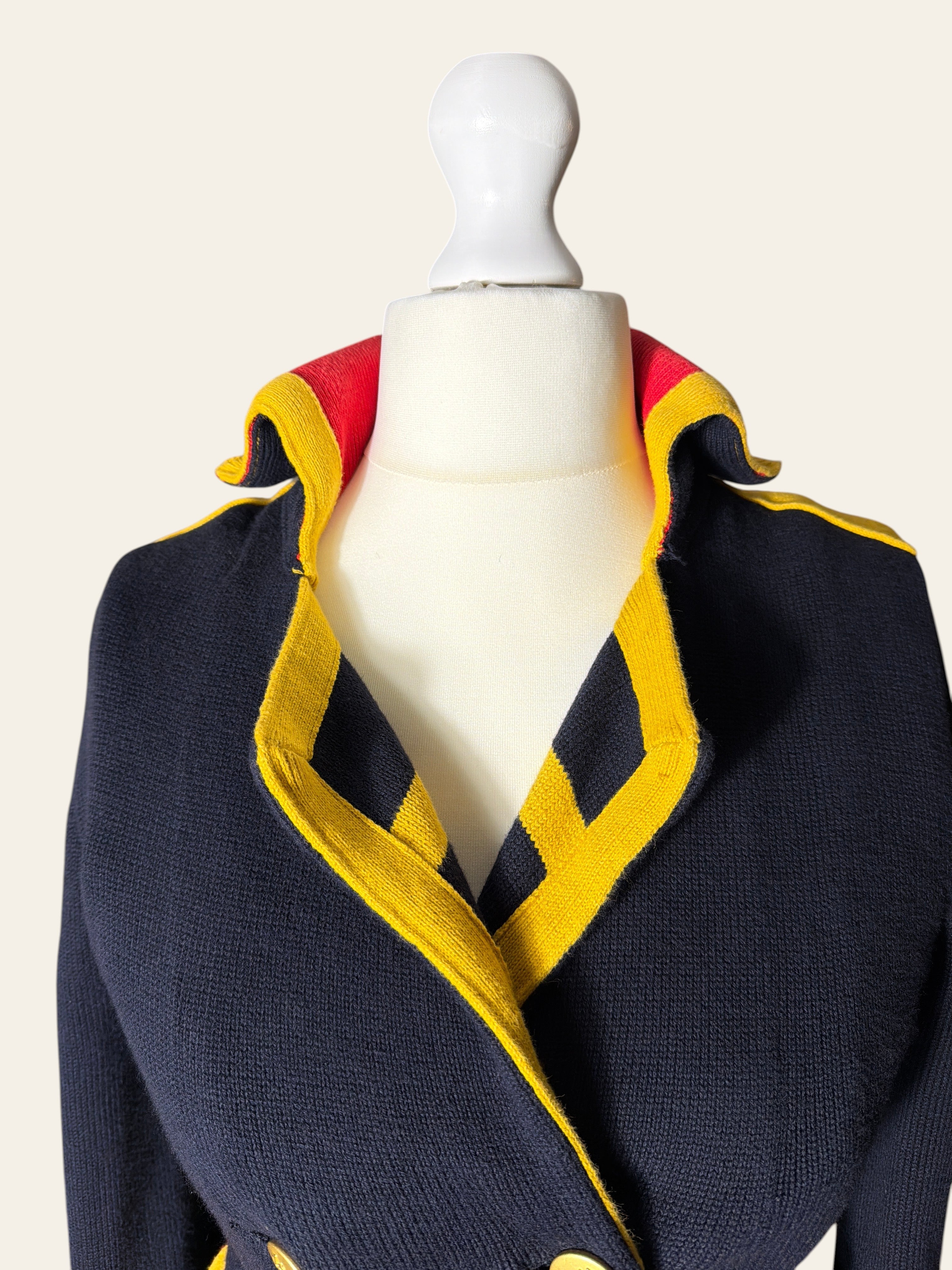 A close-up of a navy blue knit jacket displayed on a white mannequin torso. The jacket features bold yellow and red striped knit trim along the collar and shoulders.