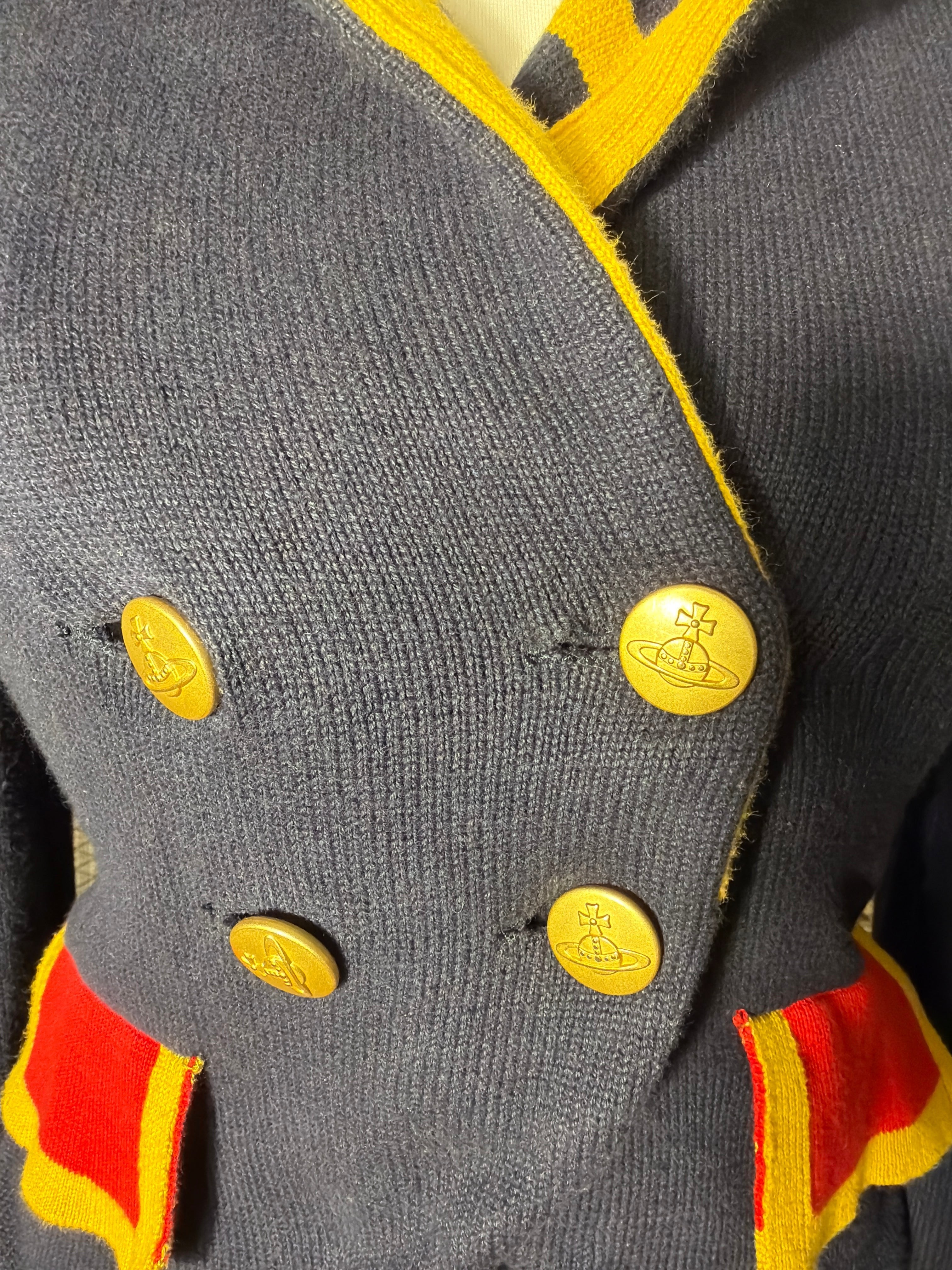 Close-up of a gray knit cardigan featuring bright yellow trim, yellow buttons with a space-themed emblem, and red and yellow striped accents near the hem.