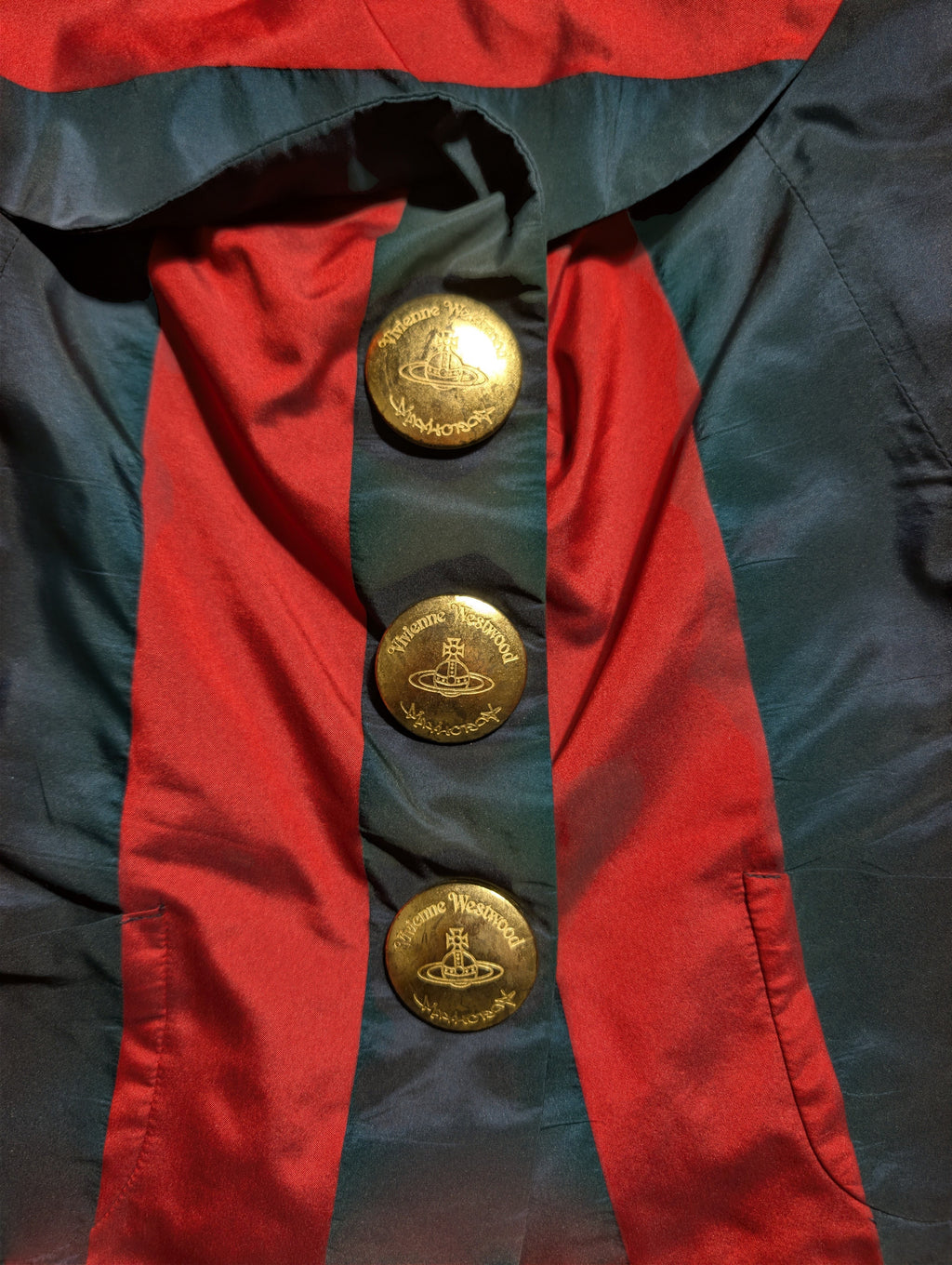 Close-up of a red and dark teal satin-like garment with three large gold buttons featuring the Orb logo, indicating the brand Vivienne Vivienne.